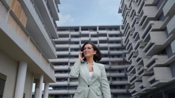 Young caucasian successful businesswoman leader wearing suit walking in big city and talking on mobile phone. Smiling woman making business call on cell on busy downtown street outdoors
