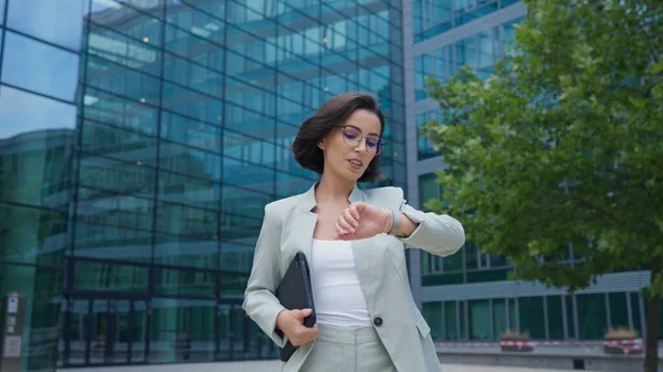 Calm businesswoman looking time on hand watch while going to the work after break. Office worker lady walking through the street in urban city district in morning hour