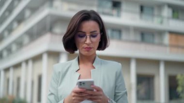 Portrait view of the happy woman looking into mobile phone screen while chatting online in city street. Young business lady smiling answering message using gadget device while looking at the camera