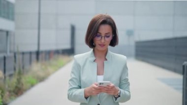 Charming positive young woman looking at the camera with smartphone on the city street. Businesswoman texting on the way to the office. Brunette adult girl messaging in social networks outdoors