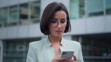 Pretty young business woman leadership chatting on smartphone, reading good messages, smiling while standing at the street in beautiful city. Modern office woman. Mobile phone