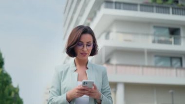 Confident business woman uses mobile phone smartphone near modern office center. Happy female employee entrepreneur worker in formal suit posing to the camera after writing text message