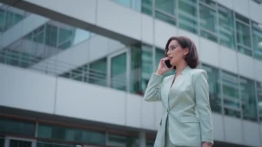 Cute businesswoman having conversation on smartphone outdoors. Portrait of beautiful caucasian female business person who talking on cell phone dressed green suit standing city street