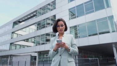 Young busy successful professional beautiful business woman holding cellphone and reading message while standing or walking on big city urban street outside during the break