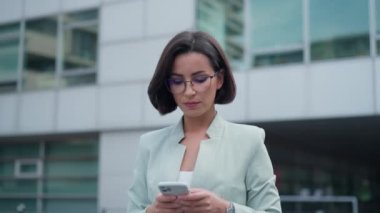 Portrait shot of elegant businesswoman standing in street on sunny day and exchanging text messages on her cell phone. Busy lady looking at the camera while tapping or scrolling on smartphone