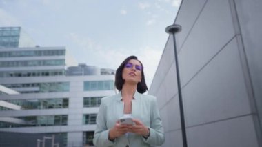 Successful businesswoman standing near office modern business center. Happy woman smiling while using smartphone, writing text message, check social media and work emails