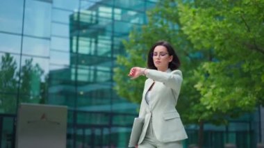 Calm businesswoman looking time on hand watch while going to the work after break. Office worker lady walking through the street in urban city district in morning hour