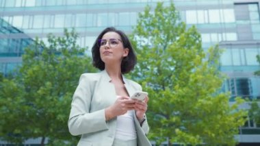 Serious businesswoman standing near office modern business center and writing message at her smartphone. Lady boss waiting for colleague