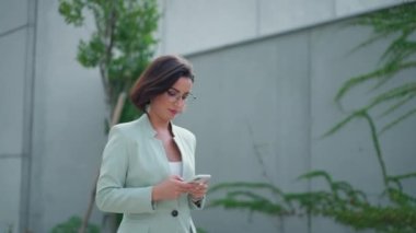 Active positive lady boss walking through the street and messaging at her phone with colleagues. Confident woman manager in formal wear taking a break and using mobile