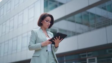 Pretty happy caucasian young woman in glasses having video chat on tablet device outdoor at business building. Joyful beautiful female talking and video chatting via webcam on gadget computer