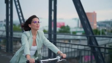 Young casual happy businesswoman enjoys riding bike in city on bridge with beautiful cityscape in the morning. Urban cycle chic and ecological transportation concept