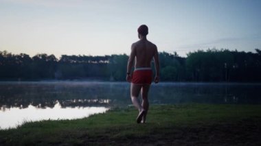 Back view of the good looking man in swim shorts going to the lake through the grass and looking at the water while preparing for the swimming in the winter morning. Ice swimming concept