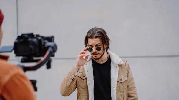 Model adam güneş gözlüklerinden bakıyor ve şehir sokağındaki kameramana poz veriyor. Çekici erkek, arka planda ateş ediyor.