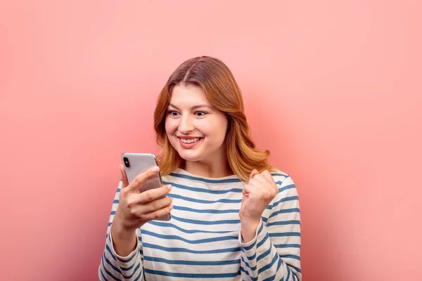 Pembe pastel arka planda izole edilmiş bir reaksiyon gibi telefon aygıtı 5G uygulama iletisi kullanan sevimli, şaşkın kahverengi saçlı kızın portresi. Mutluluk kavramı.