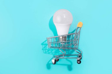 İzole edilmiş mavi arka planda, alışveriş arabasındaki lamba. Düz yatıyordu. Ekolojiyi kavra, dünyayı kurtar, fikri, enerjiyi, ekonomiyi, tasarrufu. Dünya günü. Akıllı alışveriş, satış, satın alma.