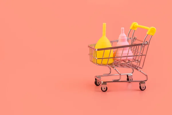 Alışveriş arabasında silikon pembe ve sarı aybaşı bardağı. Pembe arka planda kadın sağlığı ve alternatif hijyen. Çevre dostu, kadın hijyeni için alternatif yeniden kullanılabilir ürün. Minimalizm organik.