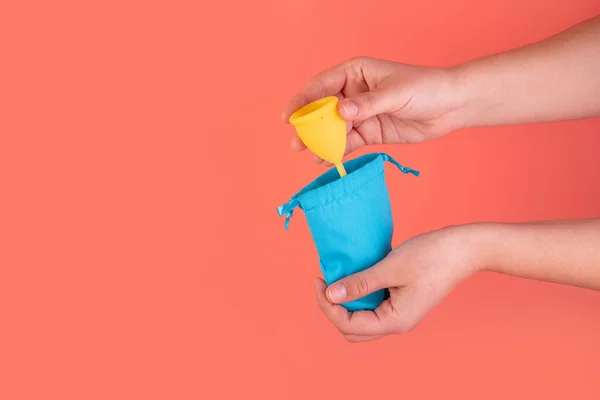 Silikon sarısı regl bardağı ve kadın eli mavi kese. Pembe arka planda kadın sağlığı ve alternatif hijyen. Çevre dostu, kadın hijyeni için alternatif yeniden kullanılabilir ürün.