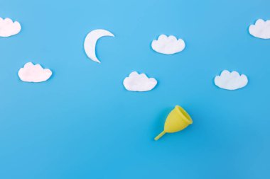 Mavi arka planda ay ve bulutların olduğu silikon adet bardağı. Kadın sağlığı ve alternatif hijyen. Çevre dostu, kadın hijyeni için alternatif yeniden kullanılabilir ürün. Yumuşak gece kavramı.