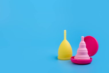Silikon pembe ve sarı aybaşı bardakları. Mavi arka planda kadın sağlığı ve alternatif hijyen. Çevre dostu, kadın hijyeni için alternatif yeniden kullanılabilir ürün. Minimalizm yumuşak, organik