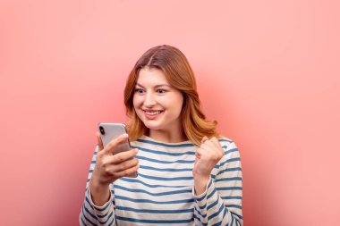 Pembe pastel arka planda izole edilmiş bir reaksiyon gibi telefon aygıtı 5G uygulama iletisi kullanan sevimli, şaşkın kahverengi saçlı kızın portresi. Mutluluk kavramı.