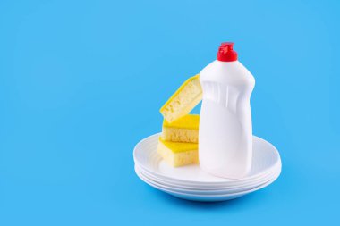 Cleaning yellow sponge, white plate and detergent for dishes on a blue background. Top view, close up. Concept cleaning company, purity, cleaning, kitche