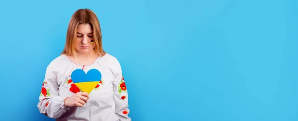 Sad young Ukrainian girl in an embroidered shirt with the flag of Ukraine on her face holds a heart of a yellow-blue flag. The concept of participation of the Ukrainian people in the war with Russia