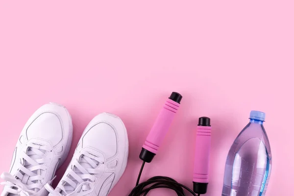 Fitness accessories concept. Top view photo of white sneakers, bottle of water and skipping, jump rope on isolated pink background and copy space. Flatlay