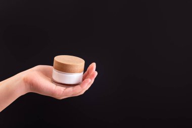 Glass jar of white moisturized cream in a female hand on black background.   Body care lotion. Simple empty packaging beauty cosmetics. Mock up, copy space
