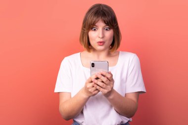 Portrait of a lovely amazed cheerful brown-haired woman using a 5g phone device as a reaction of delight and huge surprise, isolated on a pink pastel background. The concept of happiness and WOW