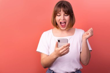 Portrait of a lovely amazed cheerful brown-haired woman using a 5g phone device as a reaction of delight and great joy, isolated on a pink pastel background. The concept of happiness.