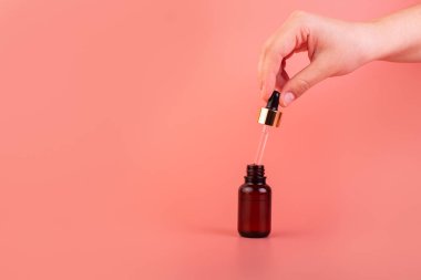 A female hand holds a dropper with a cosmetic serum. Natural cosmetic product for skin purity and hydration. Brown glass bottle with a product for women's face skincare on a pink background
