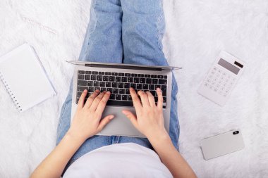Laptop, smartphone, notebook and calculator in girl's hands sitting on a soft white blanket. Flatlay, concept freelance, rest and work place