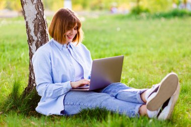 Photo portrait of pretty young woman smiling teeth sitting in green park working on laptop smiling
