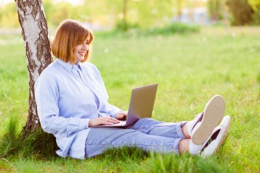 Photo of attractive lady student sit ground lean tree browsing notebook chatting friends do home tasks inspiration nature beauty harmony wear t-shirt jeans park outdoors