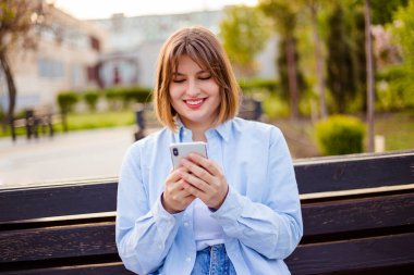 Photo of charming pretty young lady communicating modern gadget sitting bench smiling outside urban city street