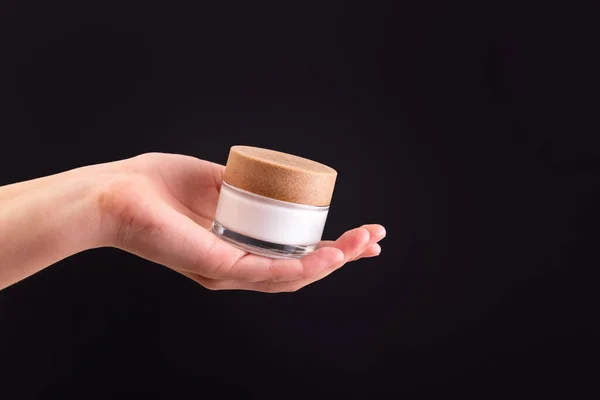 Glass jar of white moisturized cream in a female hand on black background.   Body care lotion. Simple empty packaging beauty cosmetics. Mock up, copy space