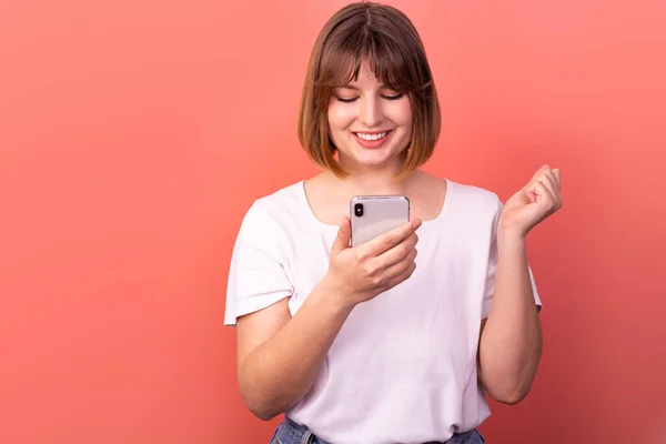 Portrait of a lovely amazed cheerful brown-haired woman using a 5g phone device as a reaction of delight and great joy, isolated on a pink pastel background. The concept of happiness.