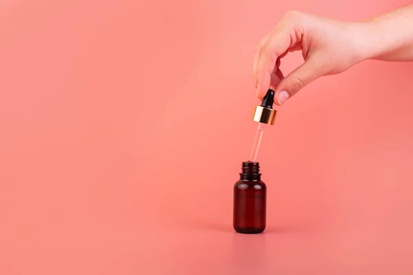 A female hand holds a dropper with a cosmetic serum. Natural cosmetic product for skin purity and hydration. Brown glass bottle with a product for women\'s face skincare on a pink background