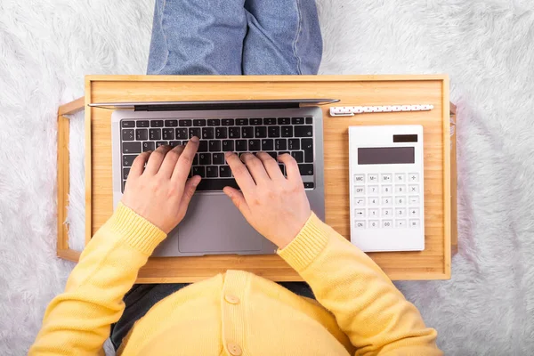 Laptop computer and calculator on tray in girl\'s hands sitting on a soft white blanket. Flatlay, concept freelance, rest and work place