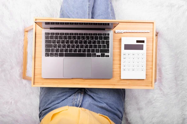 Laptop computer and calculator on tray in girl\'s hands sitting on a soft white blanket. Flatlay, concept freelance, rest and work place