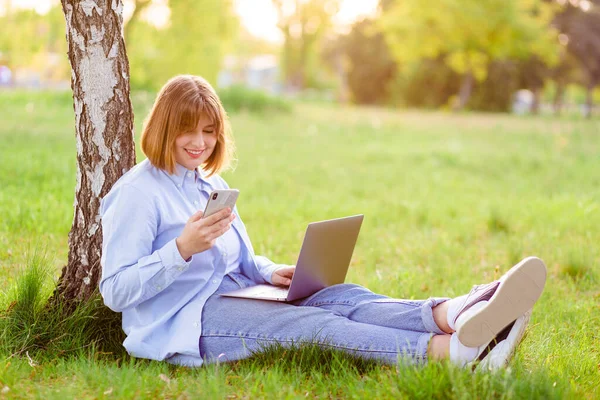 Photo portrait of a beautiful young woman smiling with her teeth, sitting in a green park and working on a laptop and looking at the phone smiling