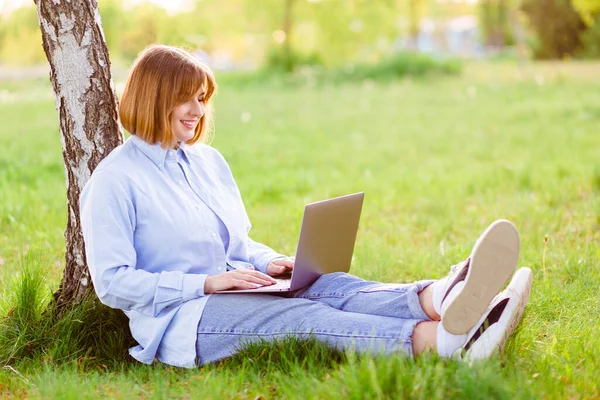 Photo of attractive lady student sit ground lean tree browsing notebook chatting friends do home tasks inspiration nature beauty harmony wear t-shirt jeans park outdoors