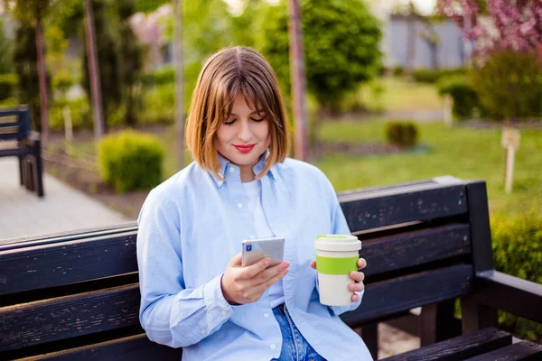 Photo of charming pretty young lady communicating modern gadget 5G with cup of tea coffee sitting bench smiling outside urban city stree