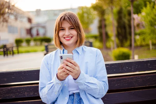 Photo of charming pretty young lady communicating modern gadget sitting bench smiling outside urban city street