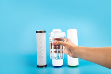 set three of cartridges for water filter with clear glass of water holding hand isolated on blue background.  Concept of water treatment technology.