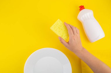Cleaning yellow sponge holding hand and detergent for dishes on a yellow background with white plates. Top view, flatlay, close up. Concept cleaning company, purity, cleaning, kitchen