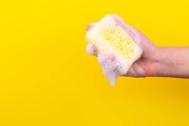Persons hand holding yellow sponge for dish wash. Washcloth covered in soap. Domestic chores and supplies concept. Sensitive dishwashing detergent. Copy space in left side. Isolated on yellow