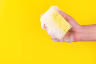 Persons hand holding yellow sponge for dish wash. Washcloth covered in soap. Domestic chores and supplies concept. Sensitive dishwashing detergent. Copy space in left side. Isolated on yellow