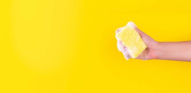 Persons hand holding yellow sponge for dish wash. Washcloth covered in soap. Domestic chores and supplies concept. Sensitive dishwashing detergent. Copy space in left side. Isolated on yellow