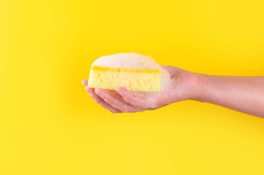 Persons hand holding yellow sponge for dish wash. Washcloth covered in soap. Domestic chores and supplies concept. Sensitive dishwashing detergent. Copy space in left side. Isolated on yellow